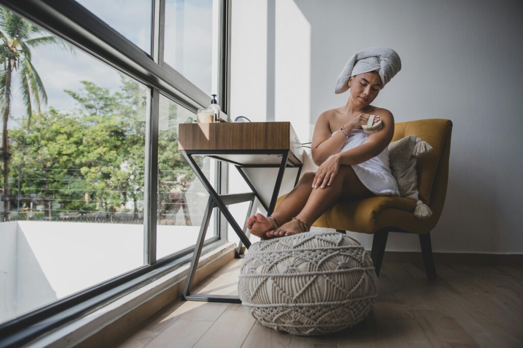 Woman enjoying a skincare routine with body scrub in a cozy, sunlit room.